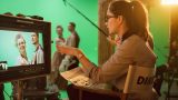 Female director sat in chair looking at a scene on a screen. Male and female actors in background