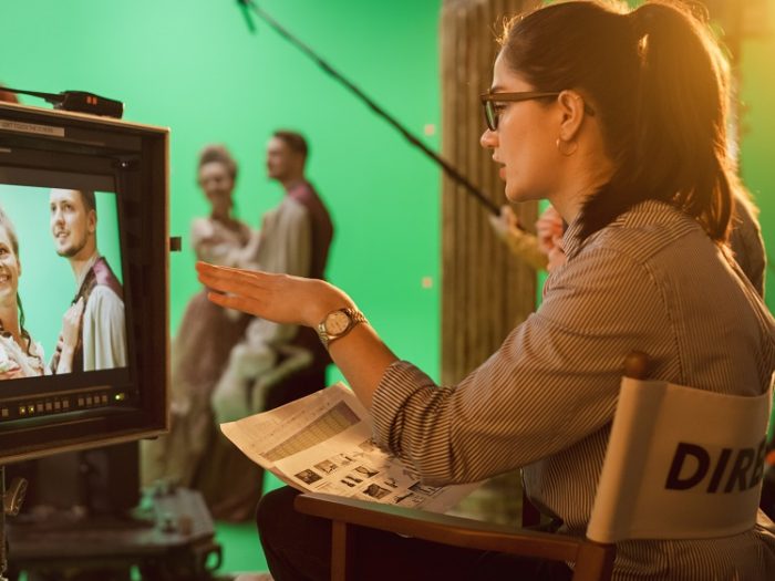Female director sat in chair looking at a scene on a screen. Male and female actors in background
