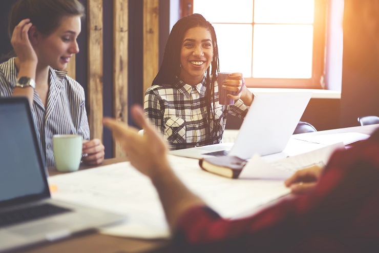business meeting with two females drinking coffee at a table with a laptop. Foreground there is an arm of another person